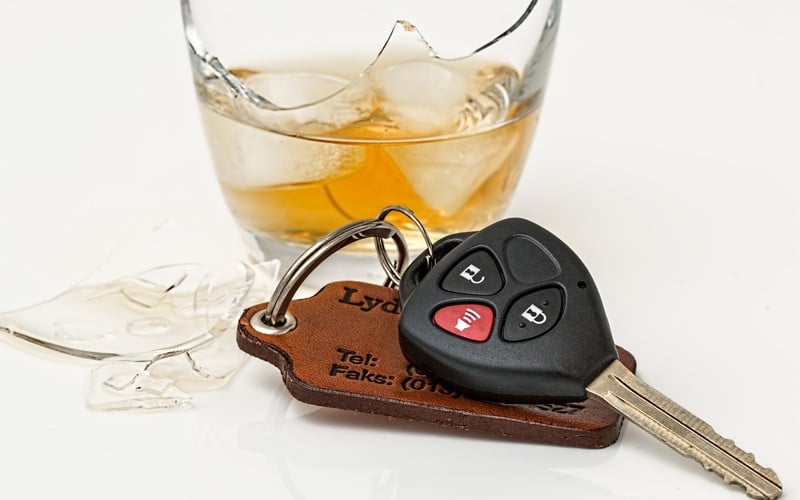 A car key with a leather fob lies next to a broken glass of whiskey with ice, symbolizing impaired driving.