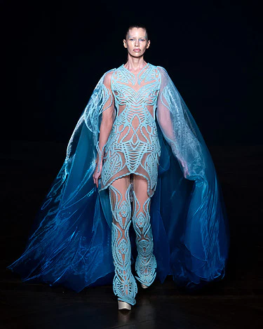 Model wearing a futuristic blue dress with intricate patterns and flowing cape on a runway, dark background.
