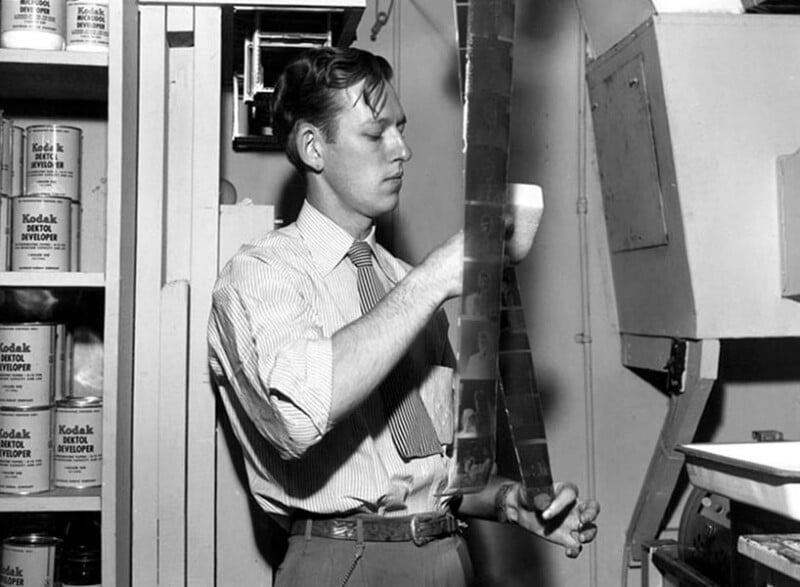 A man in a striped shirt and tie examines a long strip of photographic film in a darkroom, surrounded by Kodak developer cans and photo equipment.