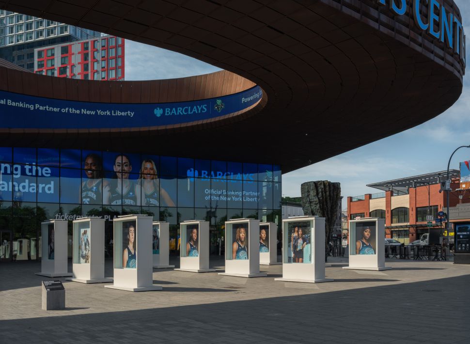 An outdoor installation of portrait photographs displayed in illuminated cases stands in front of the Barclays Center, with branding for the New York Liberty visible across the building’s façade.