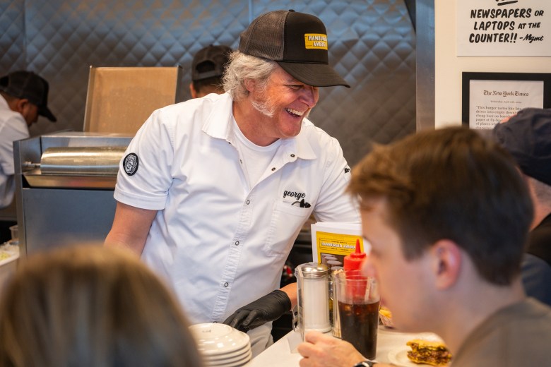 Man working in restaurant leans to his left as he smiles at patrons across the counter.