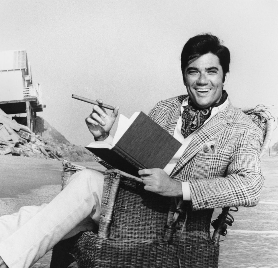Portrait of American film critic & journalist Rex Reed, in a blazer, cravat, and trousers, as he sits barefoot in a wicker chair, partially in the water, at an unspecified beach, Malibu, California, 1973. (Photo by Ellen Graham/Getty Images)