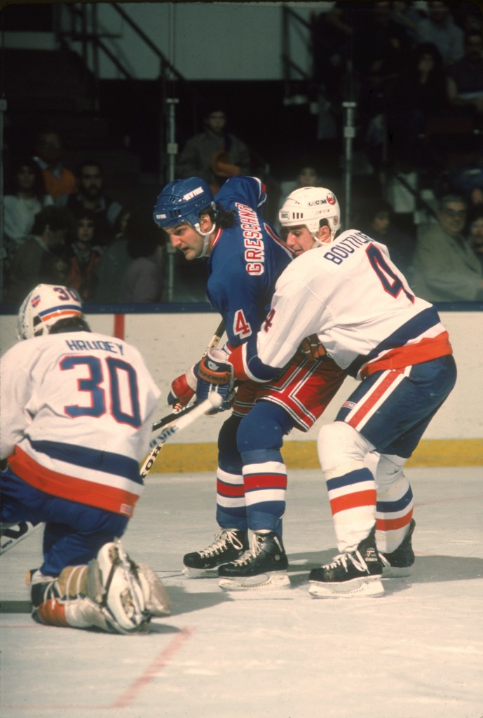 Held from behind by Canadian ice hockey player Paul Boutilier (right) of the New York Islanders, Ron Greschner (center) of the New York Rangers tries to sneak the puck past Islanders goalkeeper Kelly Hrudey during a game, November 1985