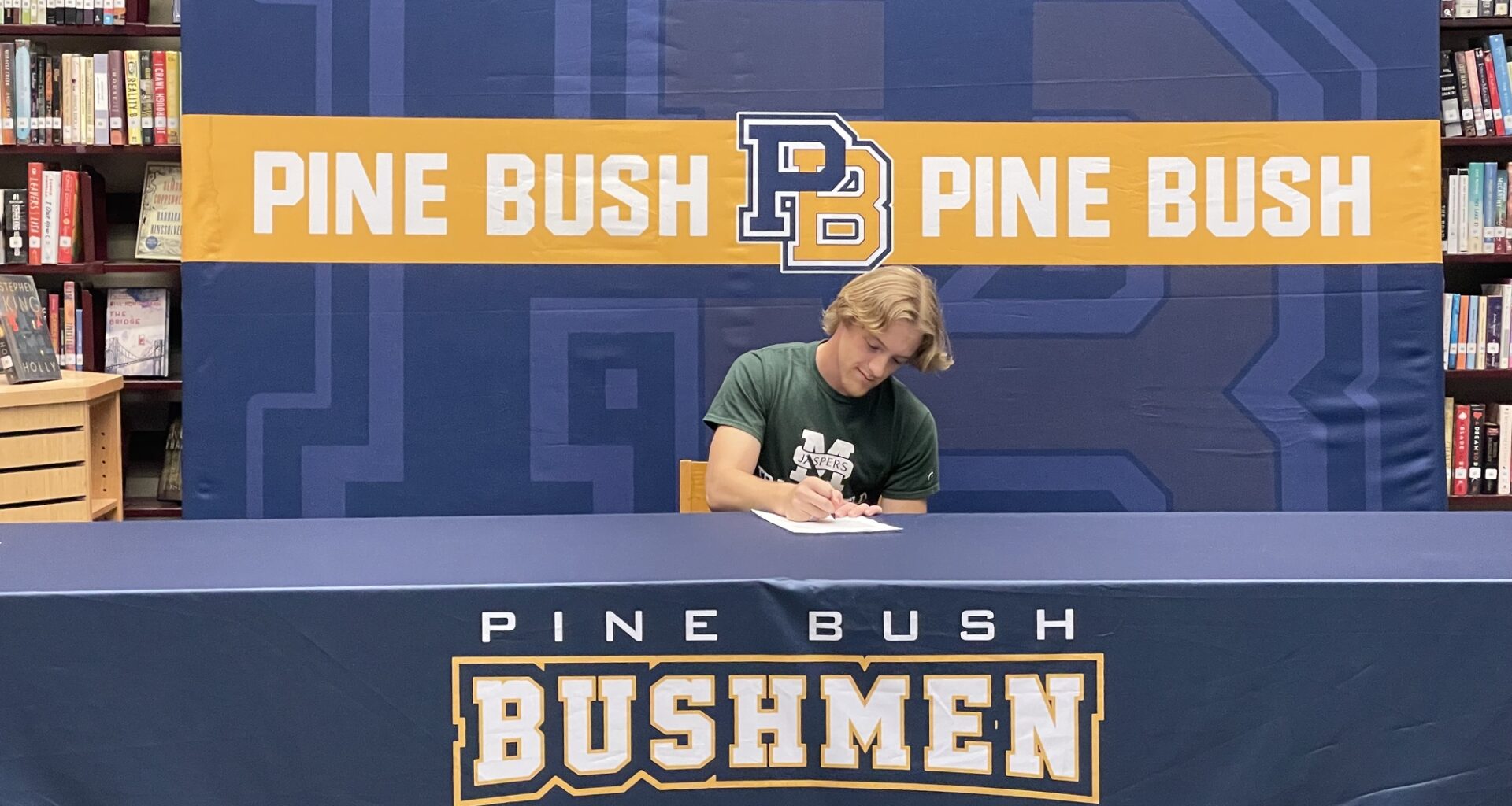 A young man wearing a green shirt with Manhattan and a large M across the front sits at a table with Pine Bush Bushmen written across the tablecloth. He signs a piece of paper.