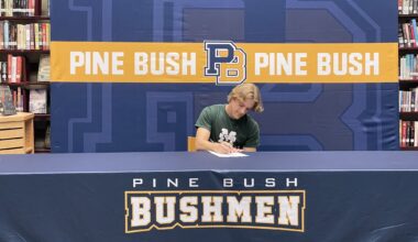 A young man wearing a green shirt with Manhattan and a large M across the front sits at a table with Pine Bush Bushmen written across the tablecloth. He signs a piece of paper.