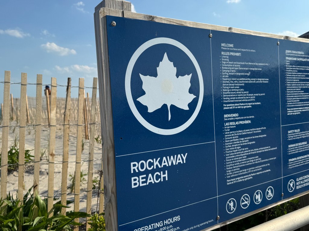 A blue Rockaway Beach sign with rules and prohibited activities, in English, Spanish, and Russian, with sand dunes and a fence in the background.