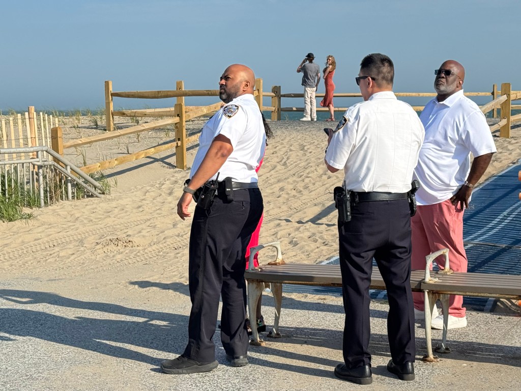 Four adults, including two uniformed officers, stand near a sandy beach with a wooden fence.