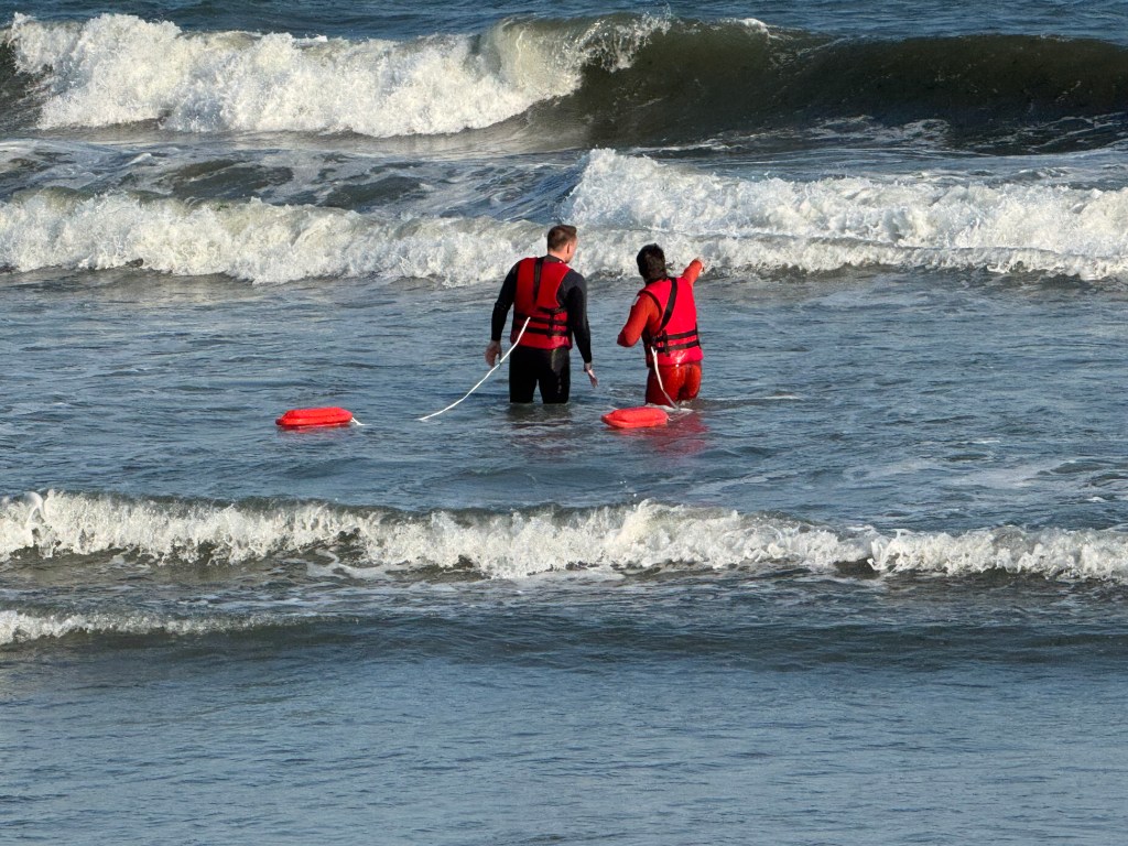 Two lifeguards in wetsuits and red life vests stand in the ocean with two rescue tubes.