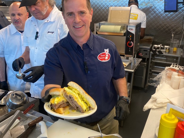 A man displays a burger at a restaurant, while others work beside or behind him.