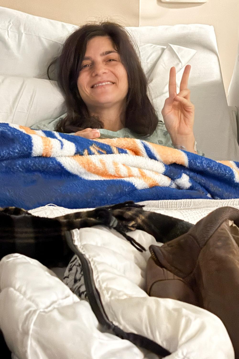 Hale lies in bed under blankets while smiling and giving the peace sign. 