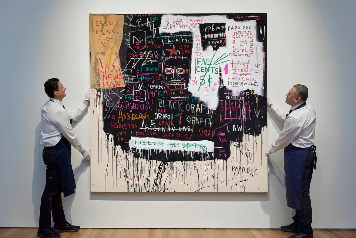 Two men in aprons adjusting a large abstract Basquiat painting to be auctioned at Sotheby's for marquee nyc sales week, painting has various words, numbers, and symbols on a white gallery wall.
