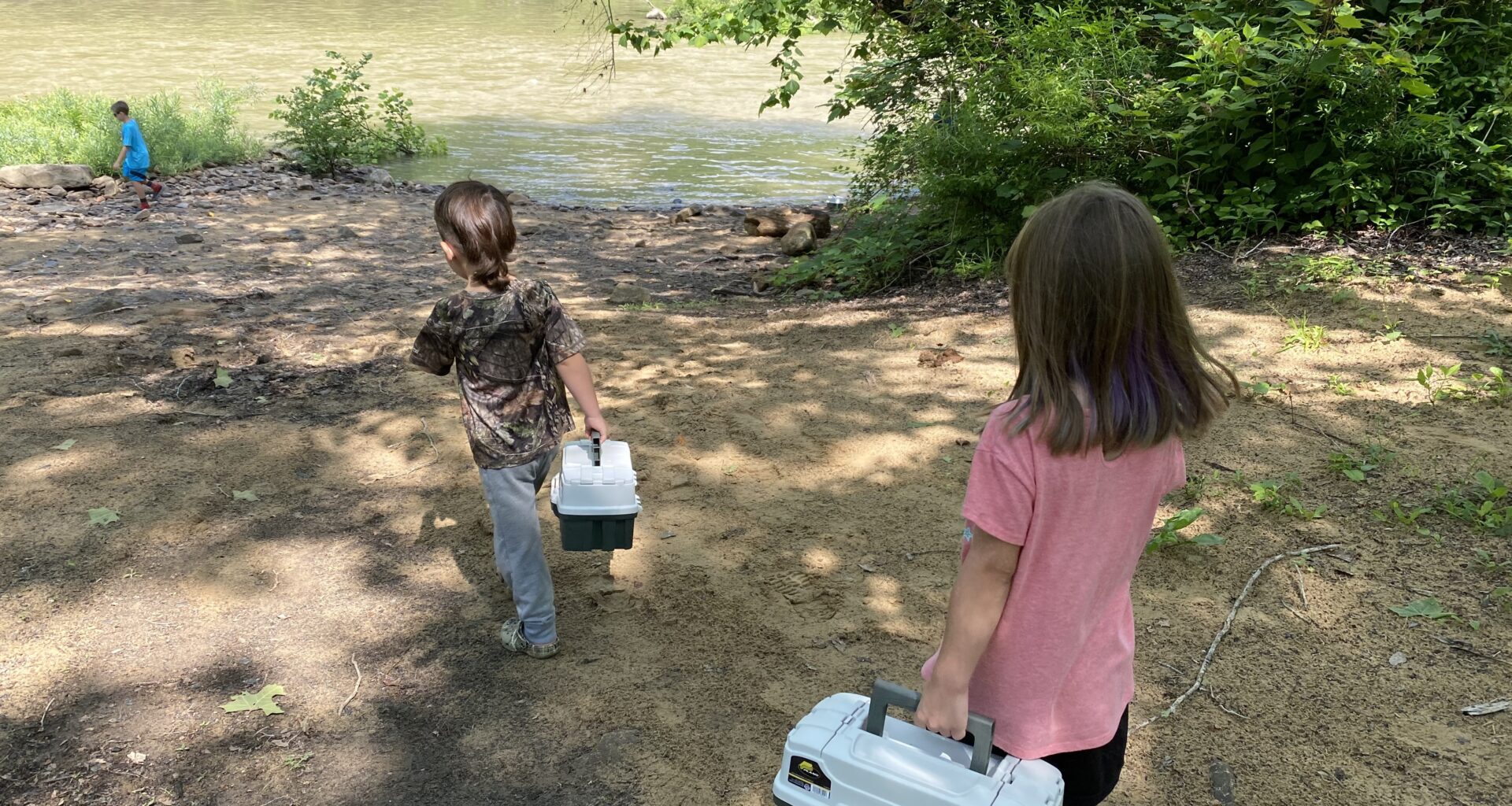 two children carrying tackle boxes walk toward a river