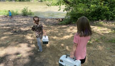 two children carrying tackle boxes walk toward a river