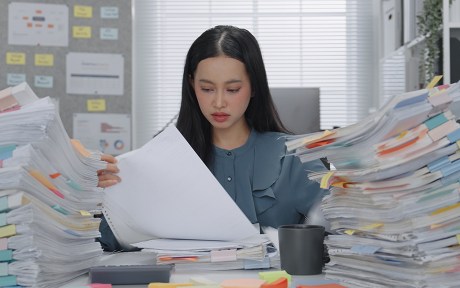 Asian Business woman working busy with massive stacks of documents check and review preparation data report or organizing paperwork workplace stress and administrative overload