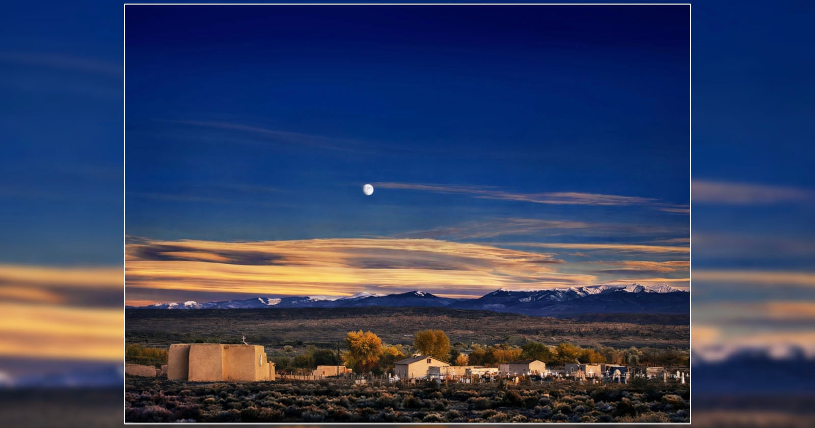 Adobe church and graveyard under a twilight sky with a full moon and snow-capped mountains in the distance.
