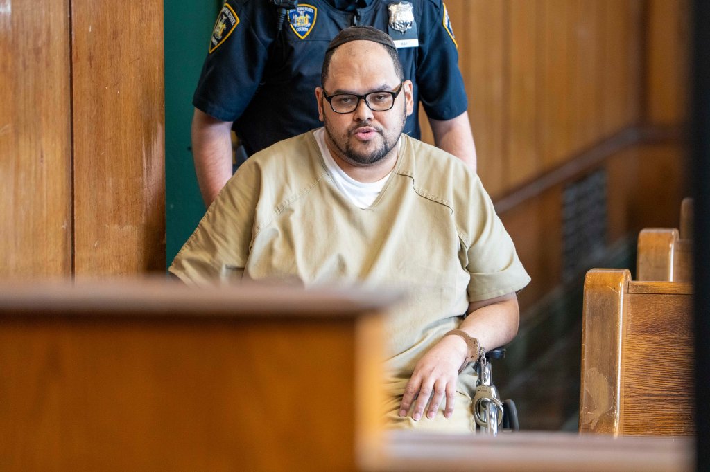 A man in an orange jumpsuit, wearing glasses and a yarmulke, sits in a wheelchair, handcuffed, with a police officer standing behind him.