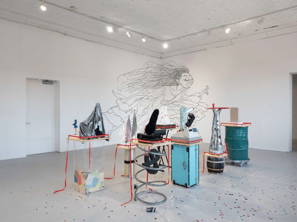 A mixed-media installation of sculptural objects, machines and colorful materials sits beneath a large wall drawing of a running figure, with confetti scattered across the floor.
