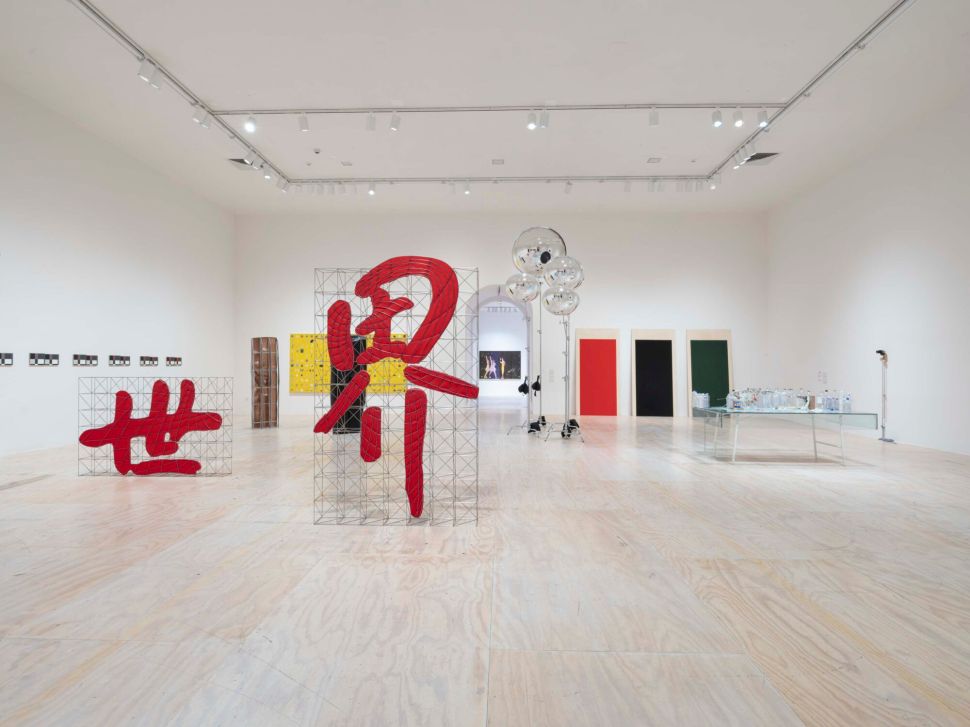 A wide gallery view shows large red abstract forms mounted on metal grids, surrounded by paintings, reflective spheres and a table of glass objects in a bright white exhibition space.