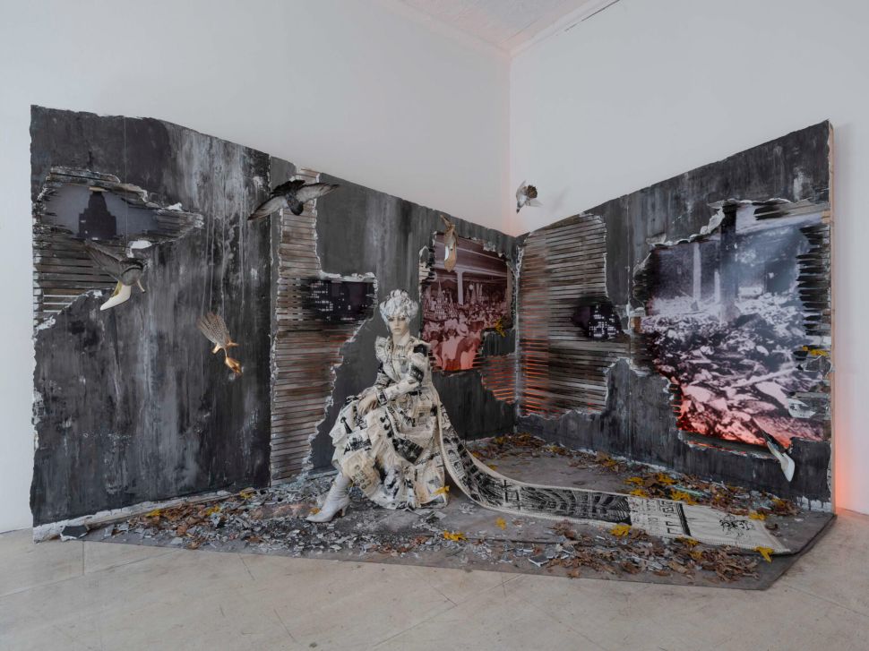 An installation features a seated figure dressed in newspaper-like fabric inside a charred, broken wall structure, with birds in mid-flight and photographic images embedded in the surfaces.