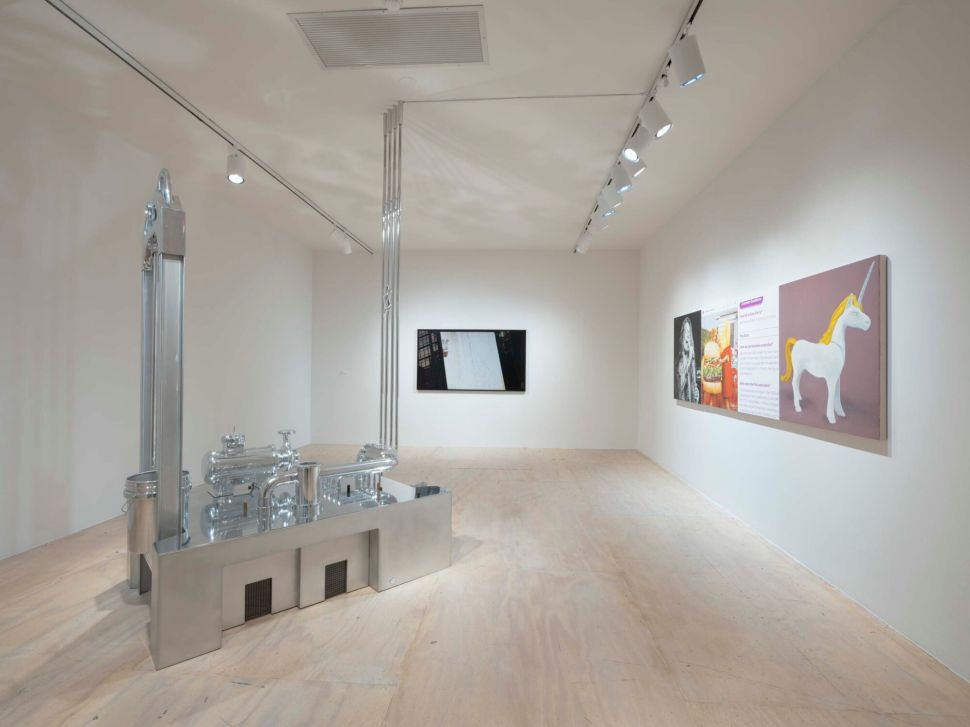 A minimalist gallery room presents a reflective metal industrial sculpture in the center, with paintings and a video screen arranged along the walls.