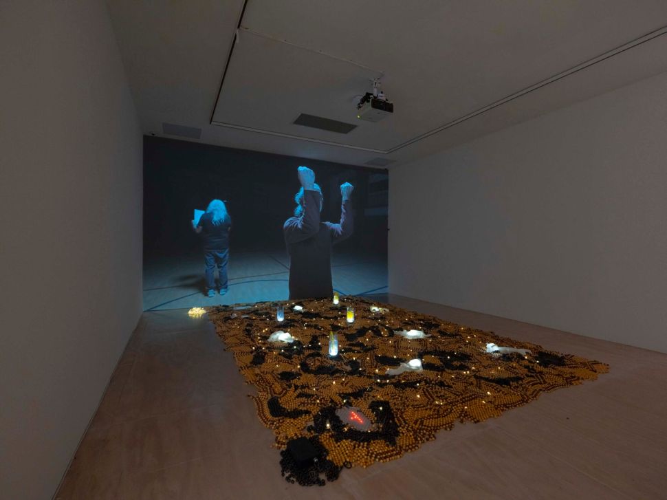 A dark gallery shows a large video projection of two figures, with an illuminated floor installation of small objects and lights spread across the room.