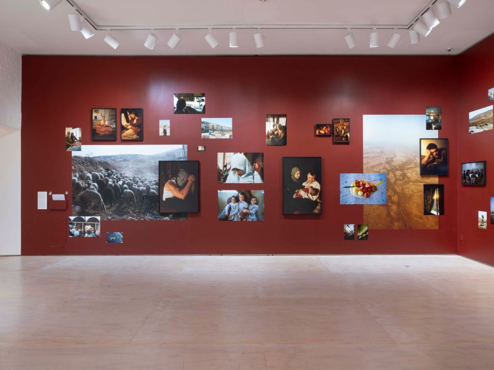 A red wall displays a salon-style arrangement of photographs of people, landscapes and everyday scenes in varying sizes across a gallery space.