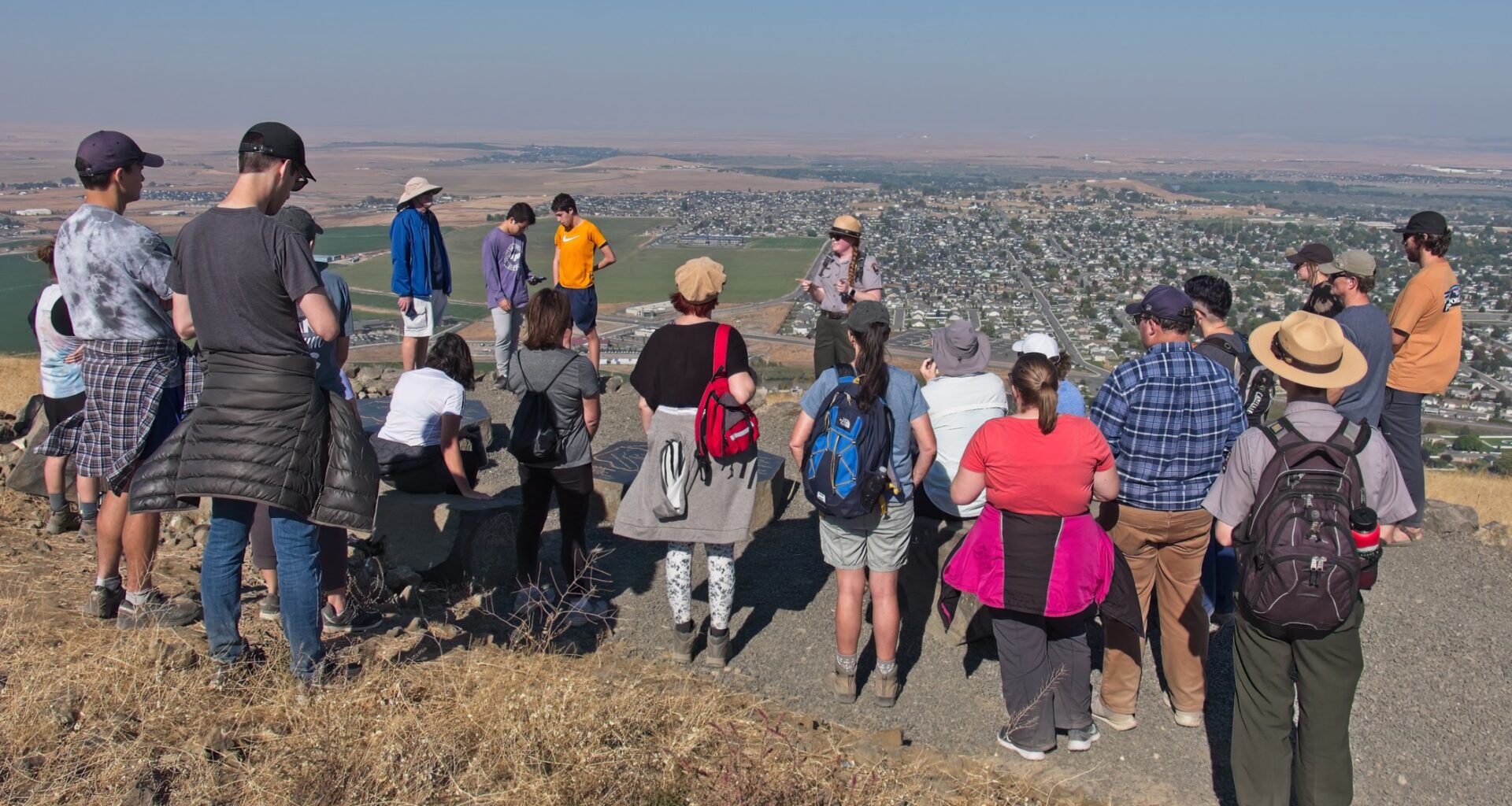 A group of about 25 people form a semi-circle around a woman addressing the crowd with expansive views of the town below on a sunny day.
