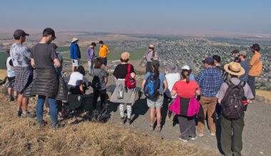 A group of about 25 people form a semi-circle around a woman addressing the crowd with expansive views of the town below on a sunny day.