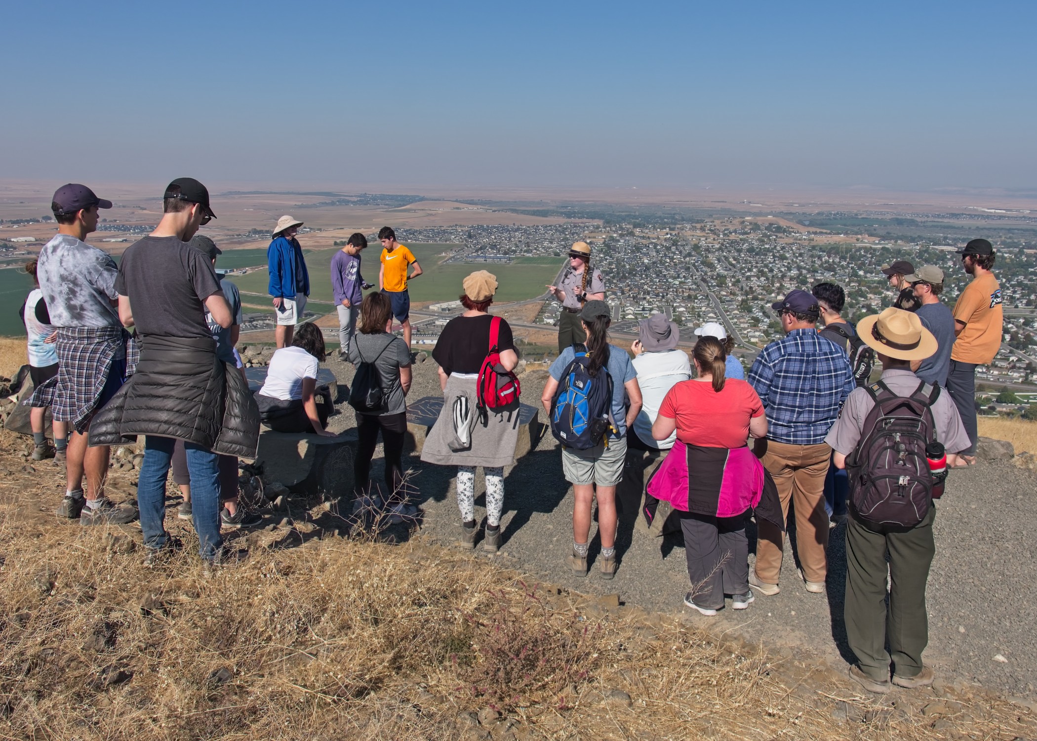 A group of about 25 people form a semi-circle around a woman addressing the crowd with expansive views of the town below on a sunny day.