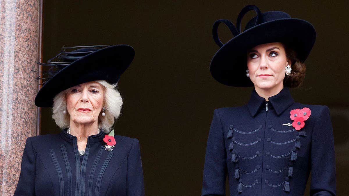 Queen Camilla and Kate Middleton in matching black attire with a red poppy pin looking serious on the balcony of Buckingham Palace.