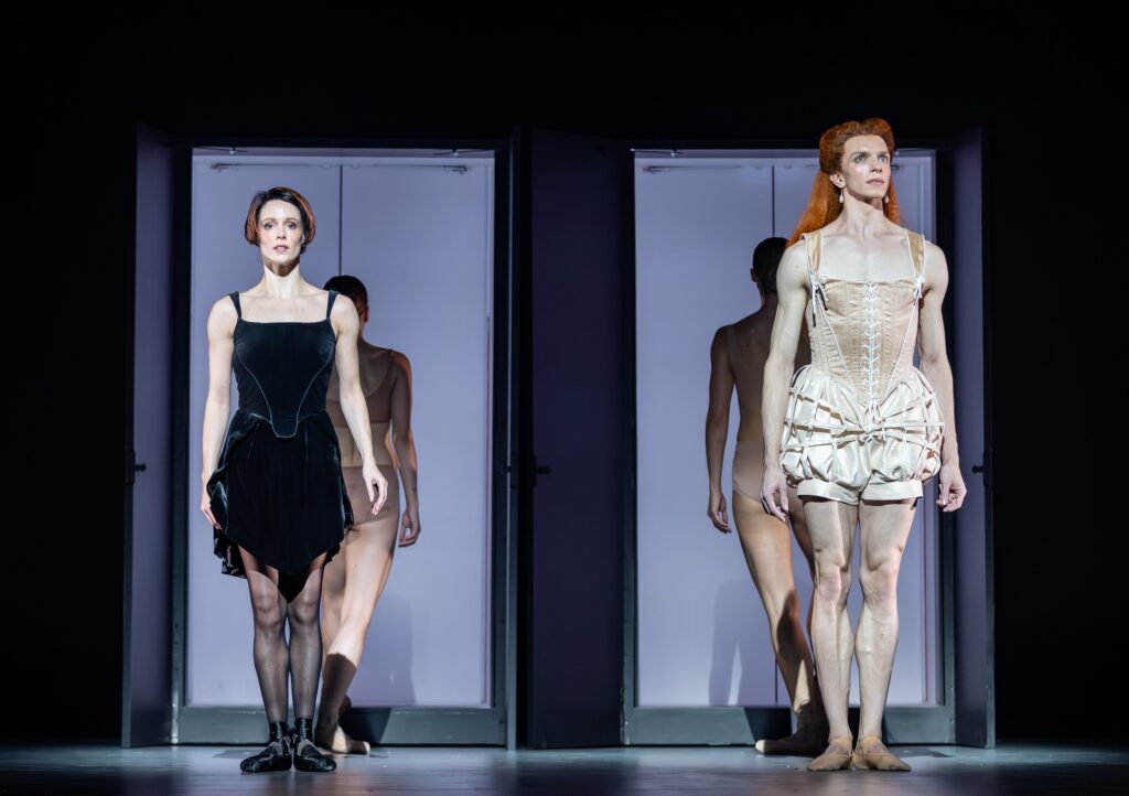 Rosanna Leney and Harvey Littelfield stand side by side with their arms by their sides, each framed by a mirror behind them. Leney, on the left, wears a black velvet, knee-length dress, sheer black tights, and black pointe shoes. Littlefield wears an off-white corset top with puffed shorts, and a long red wig.