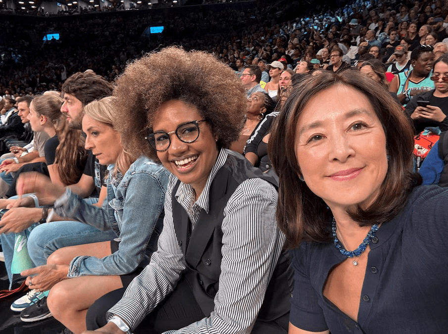 A selfie taken courtside shows two smiling women seated among a packed arena crowd, with spectators closely lined in rows behind them during a live game.