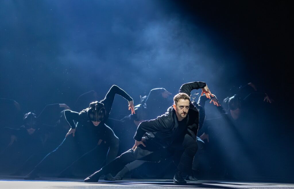 Thomas Edwards, wearing a dark suit, lunges low to the ground with his right leg stretched out to the side. He lifts his curved left arm and spreads his fingers wide. Dancers in dark costumes and masks make the same pose behind in on the smoke-filled stage.