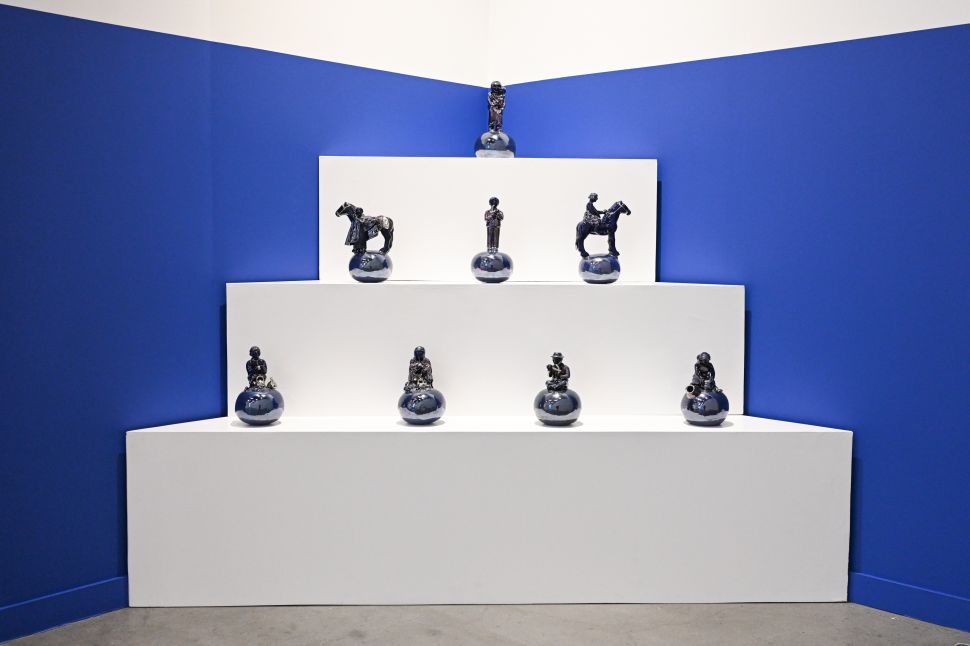 A blue-walled booth presents small glossy black ceramic sculptures arranged on tiered white plinths.