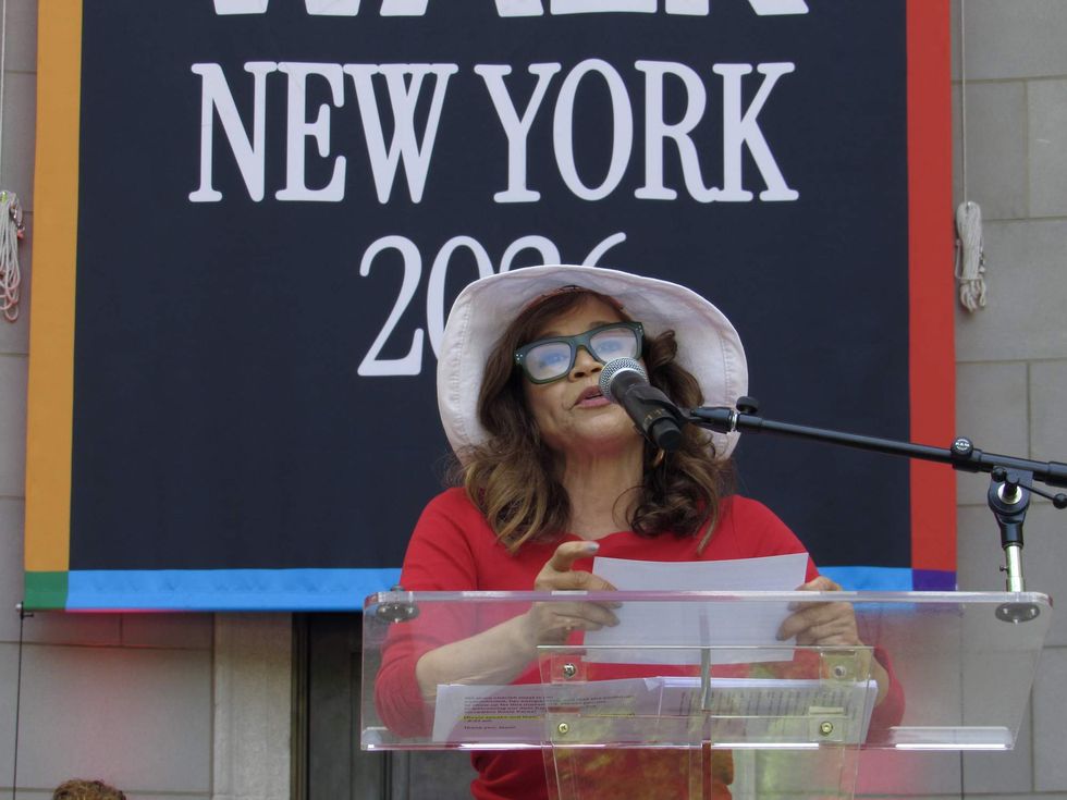 A woman in a red shirt, white brimmed hat and black glasses reads a paper into a microphone. Behind her is a banner that is partially visible, reading "New York 2026."