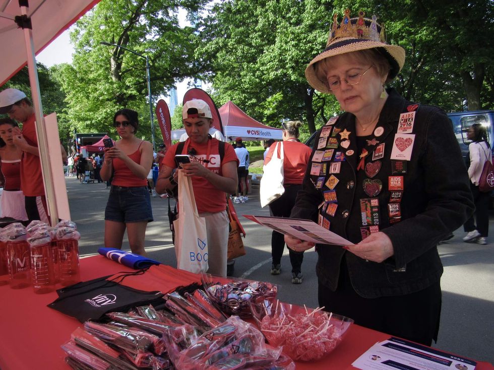 A woman wearing a hat and lots of colorful pins reads a flyer at a table with lots of merch displayed on it.