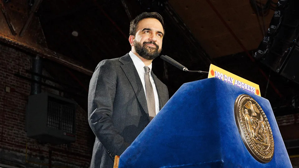 Zohran Mamdani in a suit stands at a podium labeled "100 Day Address" in a large hall with a wooden ceiling.