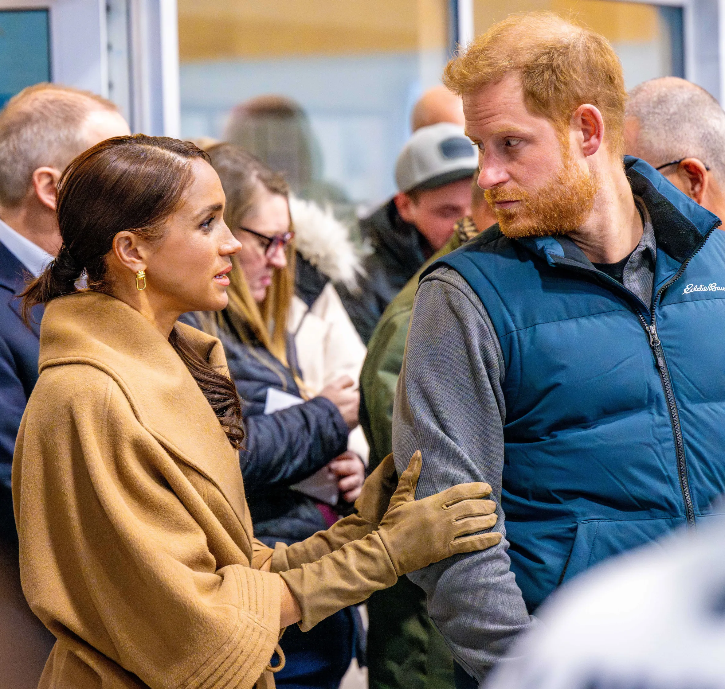 Meghan Markle, Duchess of Sussex, speaks to Prince Harry, Duke of Sussex, while wearing a camel-colored coat and gloves.