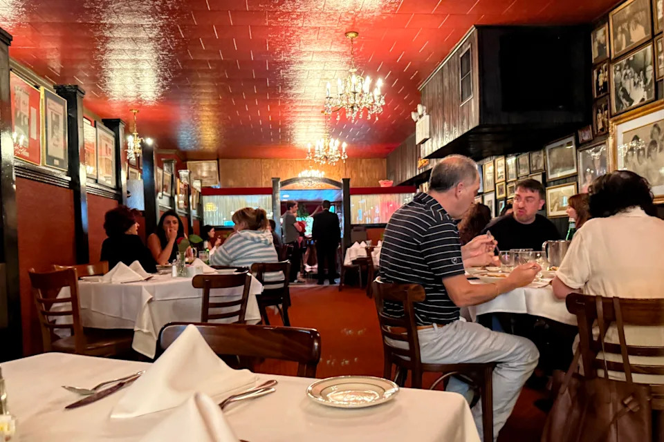 Diners are gathered at white-clothed tables in a dining room with red ceiling, red carpet, and red walls covered with photographs.
