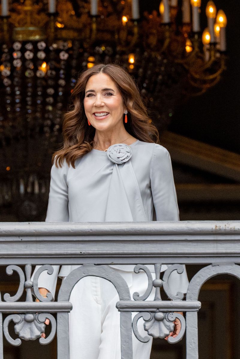 Queen Mary embraces understated elegance in a grey rosette blouse paired with crisp white tailored trousers.