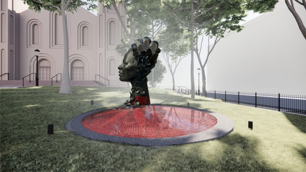 Bronze female head sculpture rises from circular red mosaic fountain beside trees and historic building façade.