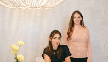 Two women sit and stand behind a table with decorative plates and a vase with yellow flowers, in front of a large chandelier.