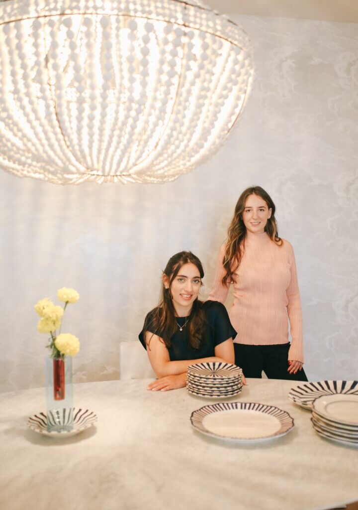 Two women sit and stand behind a table with decorative plates and a vase with yellow flowers, in front of a large chandelier.