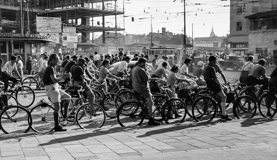 A dense mass of cyclists waits at a Munich intersection against a backdrop of postwar construction, trams and 1950s traffic.