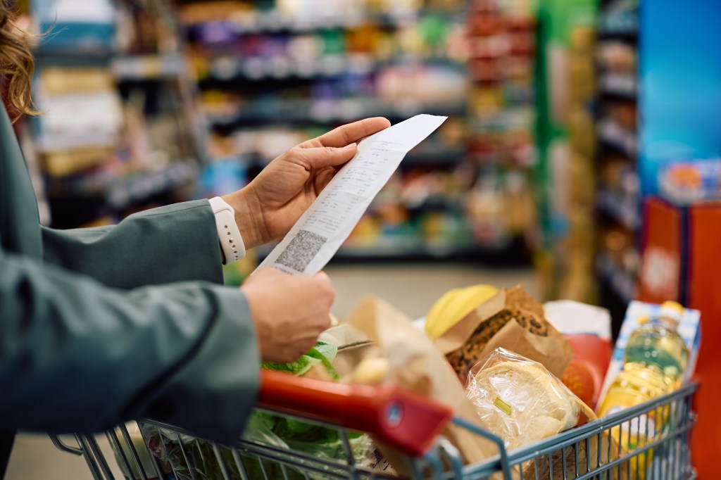 A woman checks her grocery bill while holding it over a shopping cart full of groceries.