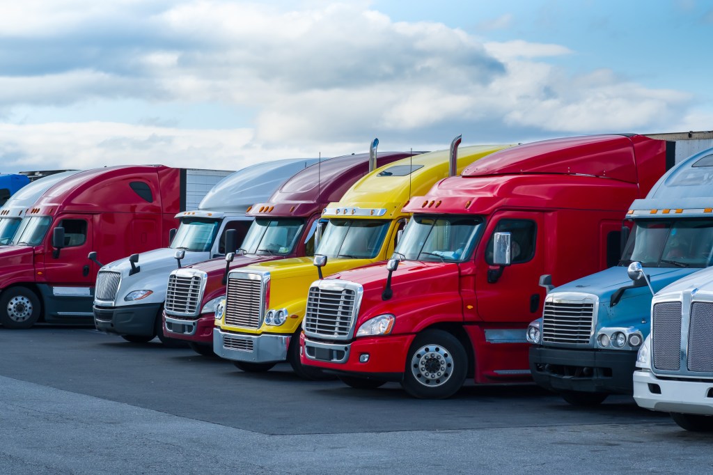 A row of colorful semi-trailer trucks parked in a lot under a cloudy sky.