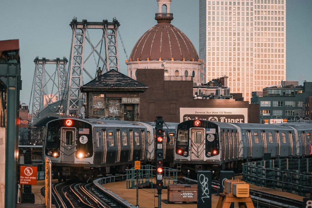 Two subway trains on elevated tracks with a bridge and a domed building in the background.