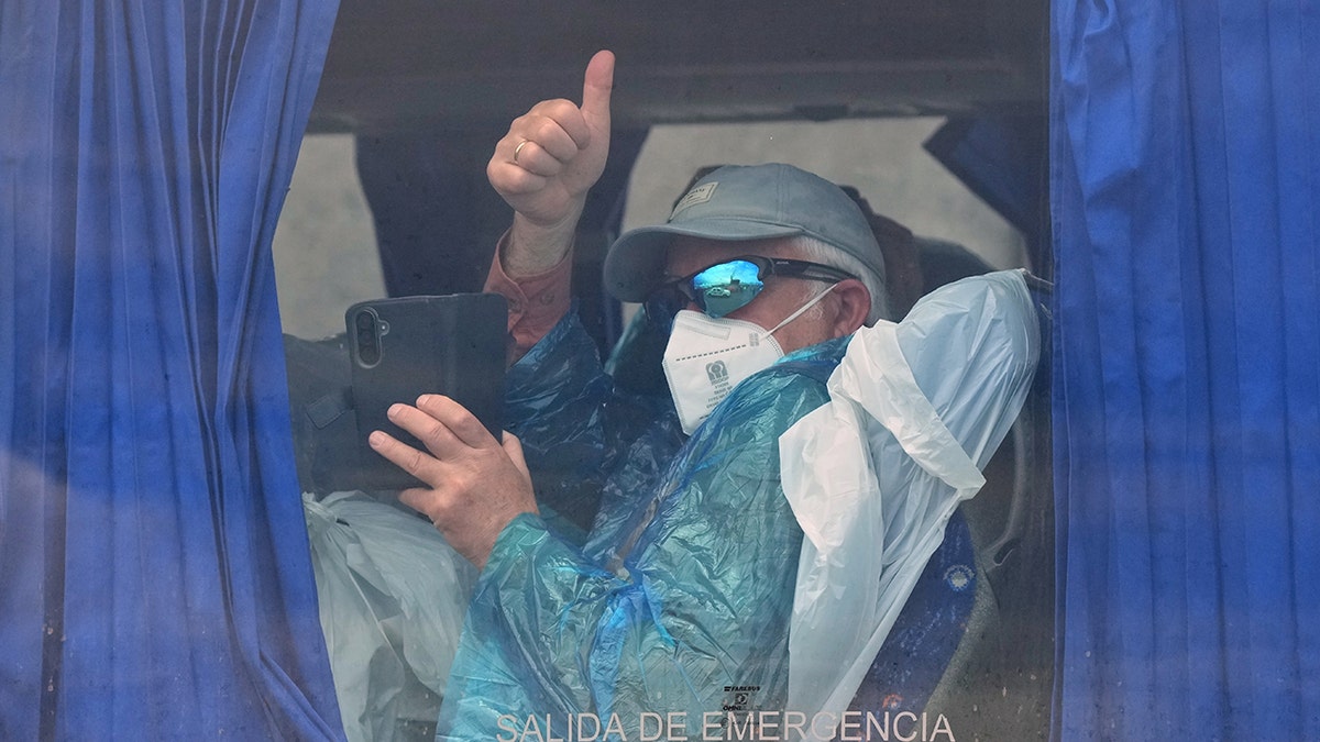 A passenger wearing a face mask and blue protective gown gives a thumbs up inside a bus.