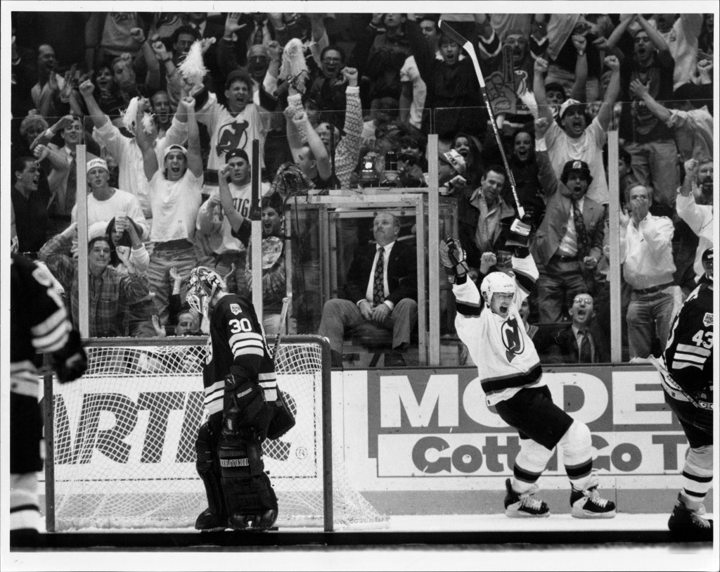Devils player Claude Lemieux celebrates a goal with his stick raised, as the crowd behind him cheers.