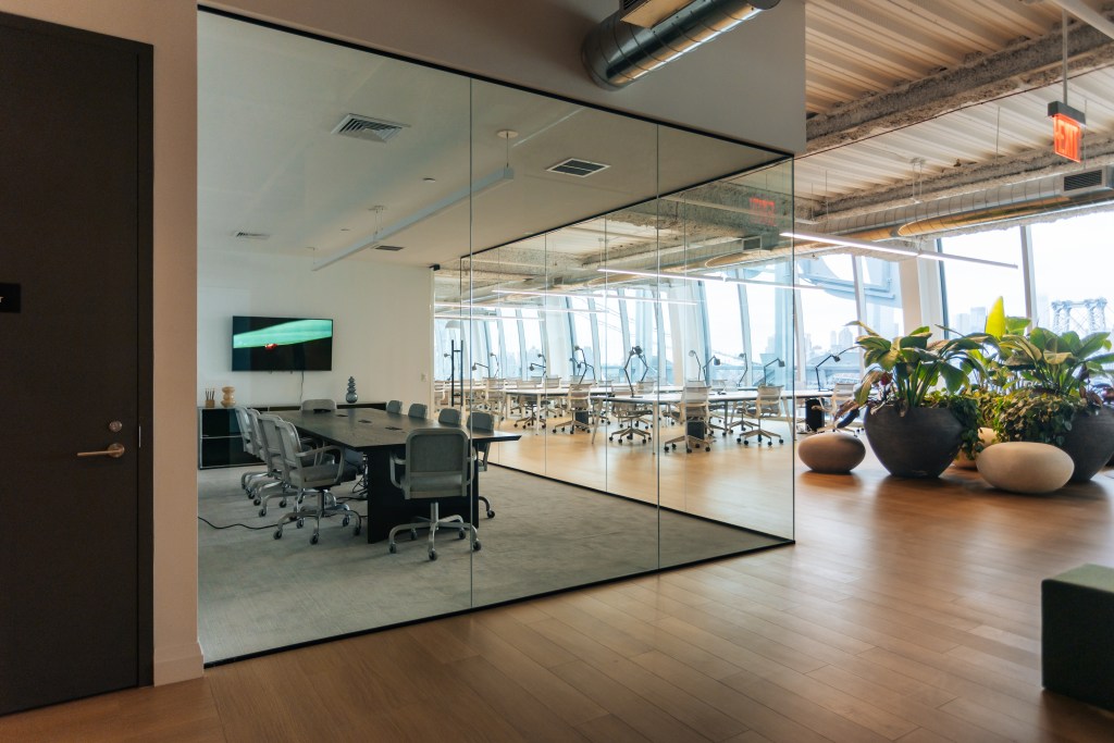 A conference room with a long table and chairs separated by a glass wall from a large office space with floor-to-ceiling windows and plants.